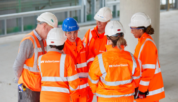 Group of six people all wearing orange PPE, stood talking/looking at each other